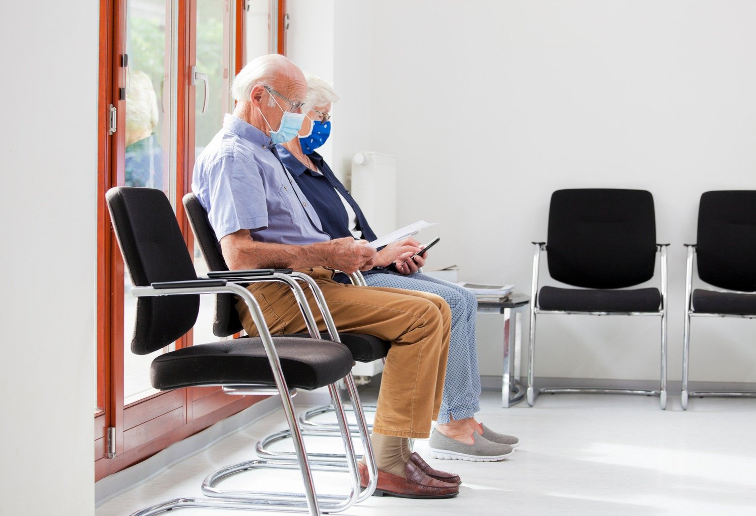 Senior-couple-sitting-with-face-masks-in-a-bright-waiting-room-of--a-hospital-or-an-office---the-man-is-reading-a-test-result---the-woman-looks-at-a-s-2